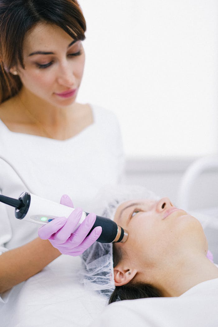 A cosmetologist uses a laser device for a facial treatment on a woman lying indoors.